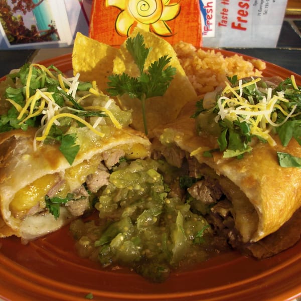 Two burritos cut in half, filled with meat and cheese, topped with salsa verde, cilantro, and shredded cheese, served with rice and tortilla chips.