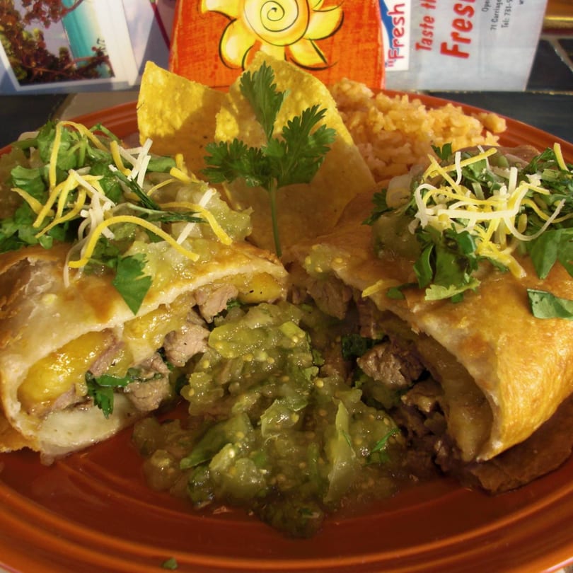 Two burritos cut in half, filled with meat and cheese, topped with salsa verde, cilantro, and shredded cheese, served with rice and tortilla chips.