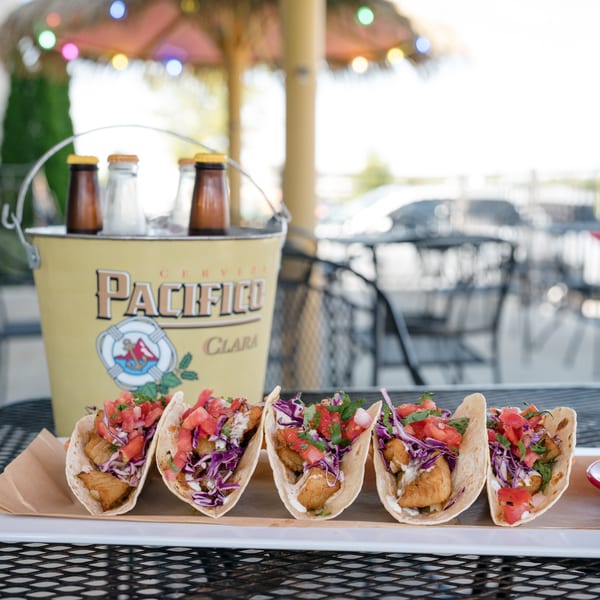 Five fish tacos with cabbage, salsa, and sauce, next to a bucket of Pacifico beer.