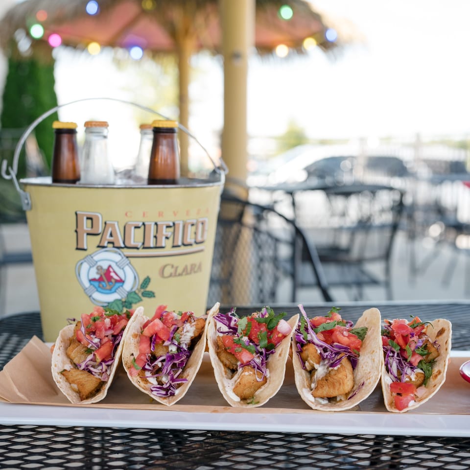Five fish tacos with cabbage, salsa, and sauce, next to a bucket of Pacifico beer.