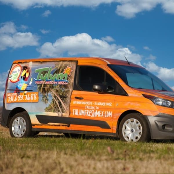 Tulum Fresh Mexican Grill's orange delivery van with tropical graphics parked on grass under a blue sky.