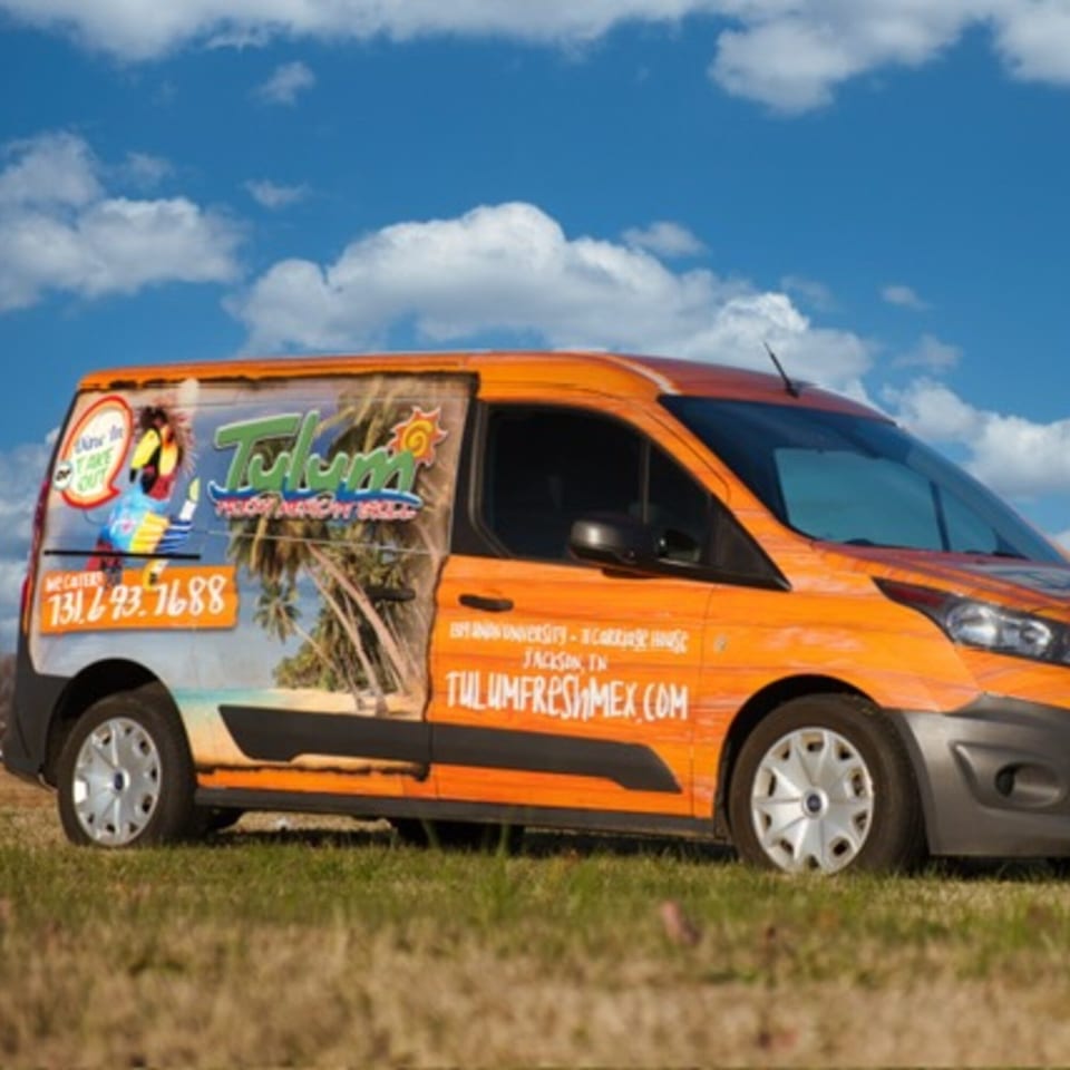 Tulum Fresh Mexican Grill's orange delivery van with tropical graphics parked on grass under a blue sky.