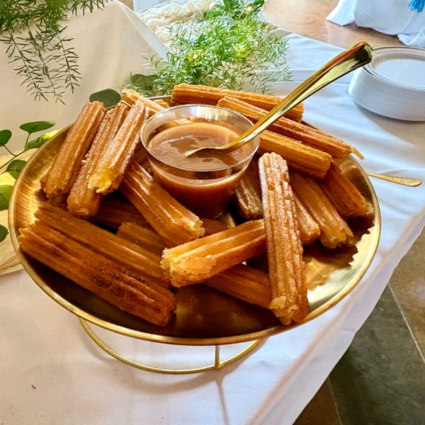 Golden churros arranged on a tiered stand with a glass of caramel dipping sauce and a gold spoon.