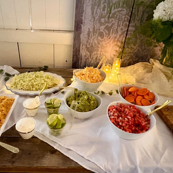 A buffet table with bowls of shredded lettuce, rice, guacamole, pico de gallo, and limes.