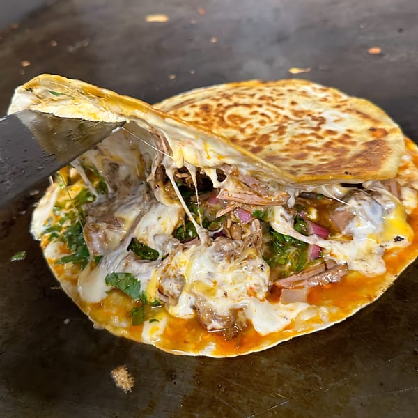 A spatula lifting a cheesy, meaty quesadilla with cilantro and red onions on a griddle.