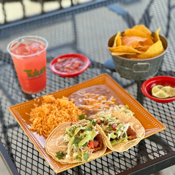 Two fish tacos with rice, beans, salsa, chips, and a pink drink on a metal patio table.
