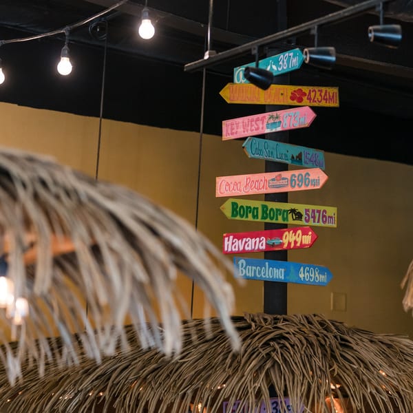 A colorful directional signpost with tropical destinations and distances, partially obscured by a thatched roof.