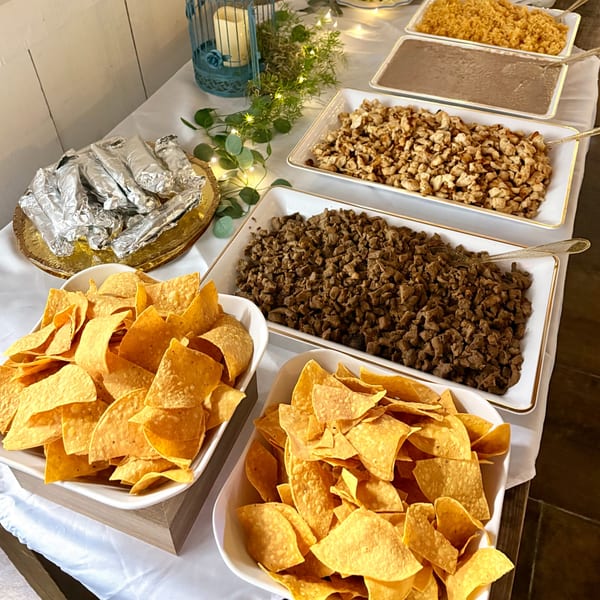 A buffet table with tortilla chips, burritos, rice, beans, and diced meats in serving dishes.