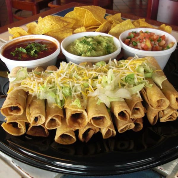 A platter of taquitos topped with shredded lettuce and cheese, served with salsa, guacamole, and tortilla chips.